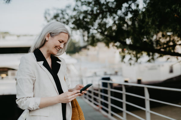 Portrait of beautiful mature woman in middle age with long gray hair, in city using her smartphone.