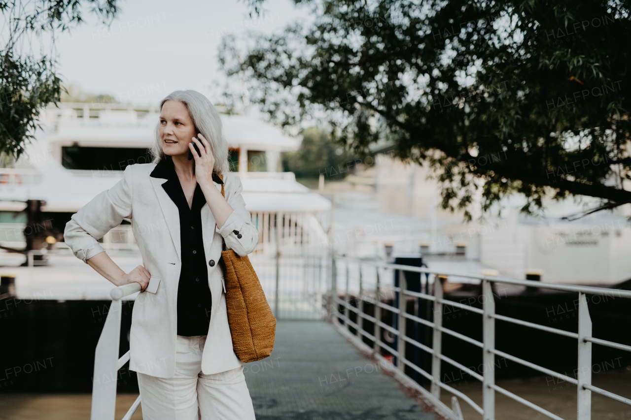 Portrait of a beautiful mature female manager with long gray hair, standing outdoors and making call. Middle-aged woman is going for walk in the city to shop.