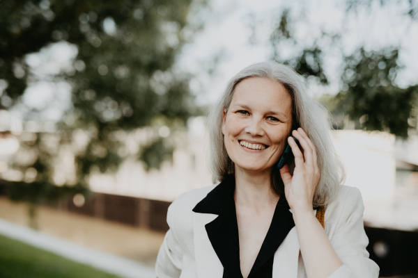 Portrait of a beautiful mature female manager with long gray hair, walking through the city. Middle-aged woman making call outdoors.