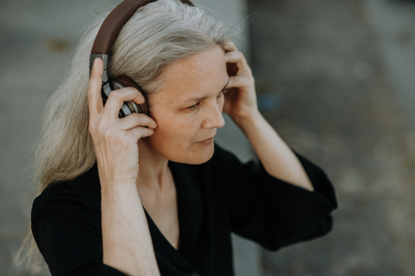 Head shot of a beautiful woman with gray hair, sitting on concrete stairs in the city, listening to music through wireless headphones.