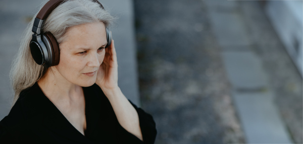 Head shot of a beautiful woman with gray hair, sitting on concrete stairs in the city, listening to music through wireless headphones. Banner with copy space.