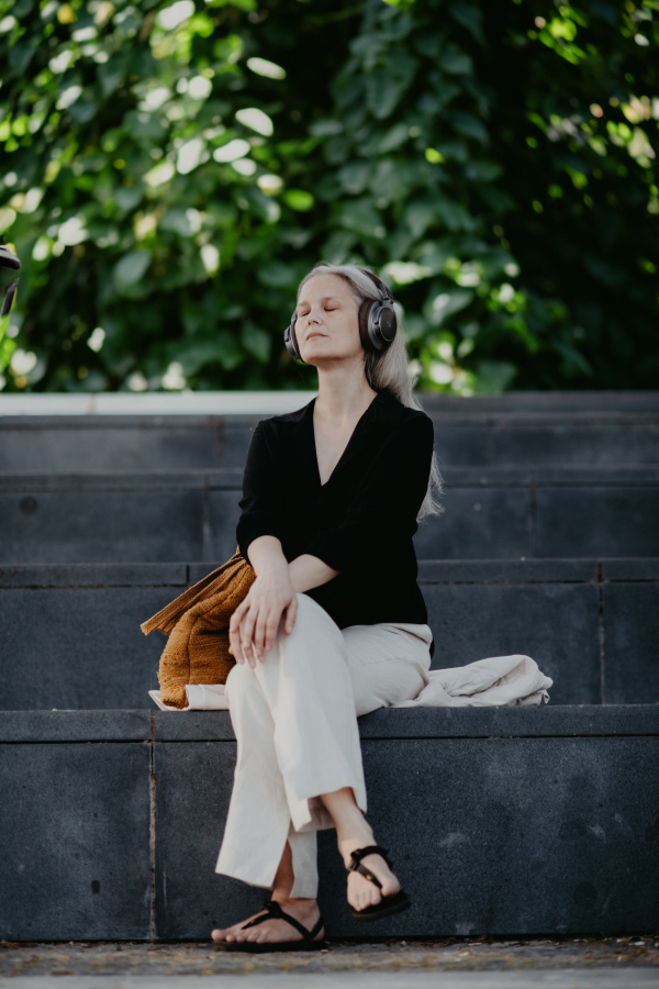 Portrait of a beautiful woman with gray hair, sitting on concrete stairs in the city, listening to music through wireless headphones.