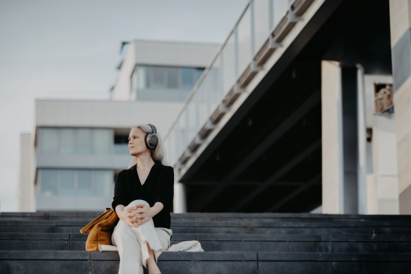 Portrait of a beautiful woman with gray hair, sitting on concrete stairs in the city, listening to music through wireless headphones. Banner with copy space.