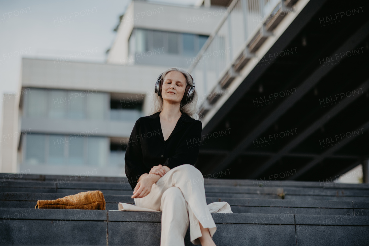 Portrait of a beautiful woman with gray hair, sitting on concrete stairs in the city, listening to music through wireless headphones.