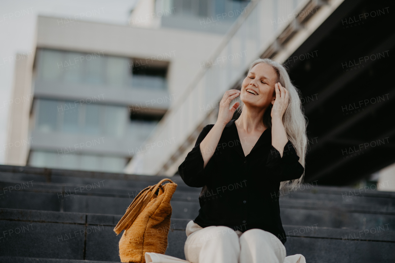 Portrait of a beautiful woman with gray hair, sitting on concrete stairs with closed eyes, enjoying warm summer weather in the city.