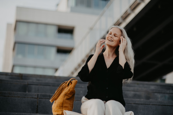 Portrait of a beautiful woman with gray hair, sitting on concrete stairs with closed eyes, enjoying warm summer weather in the city.