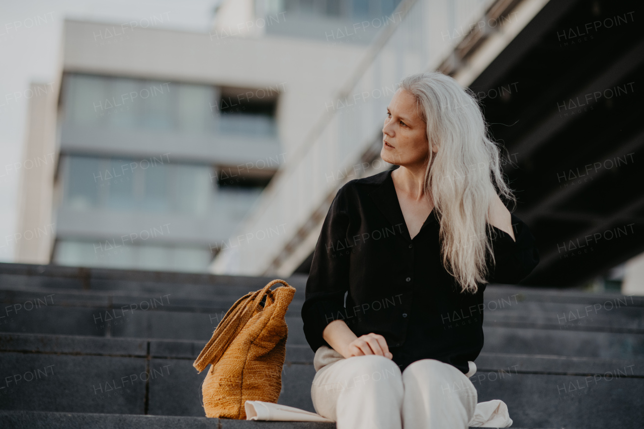 Portrait of a beautiful woman with gray hair, sitting on concrete stairs with closed eyes, enjoying warm summer weather in the city.