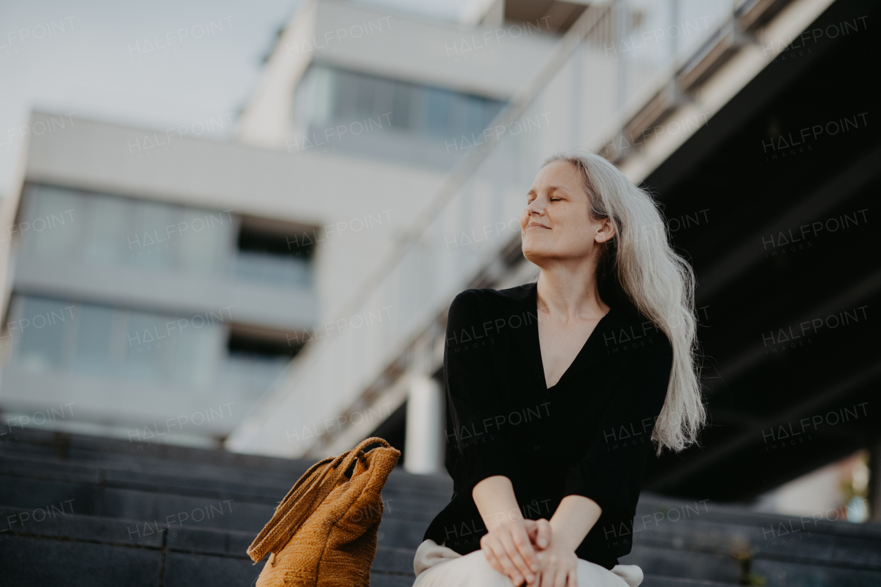 Portrait of a beautiful woman with gray hair, sitting on concrete stairs with closed eyes, enjoying warm summer weather in the city.