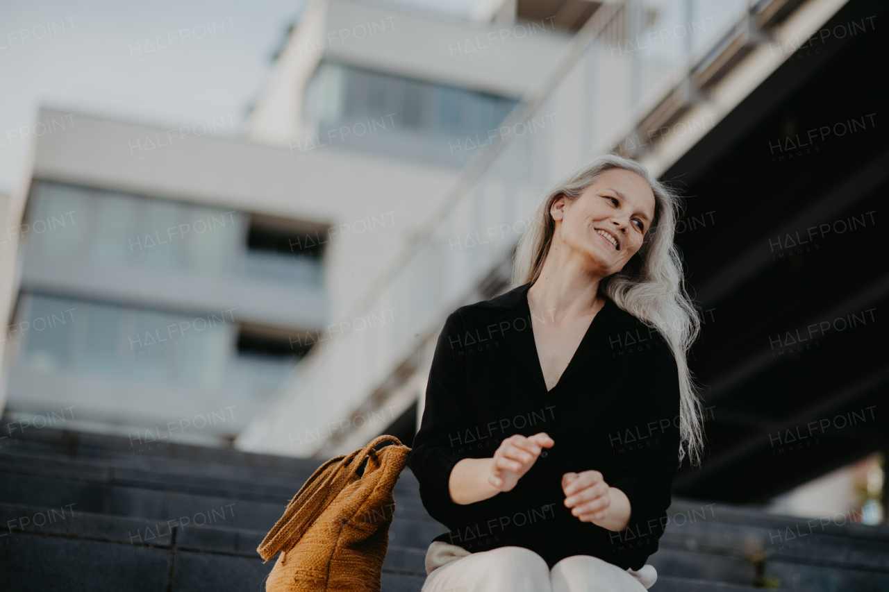 Portrait of a beautiful woman with gray hair, sitting on concrete stairs with closed eyes, enjoying warm summer weather in the city.