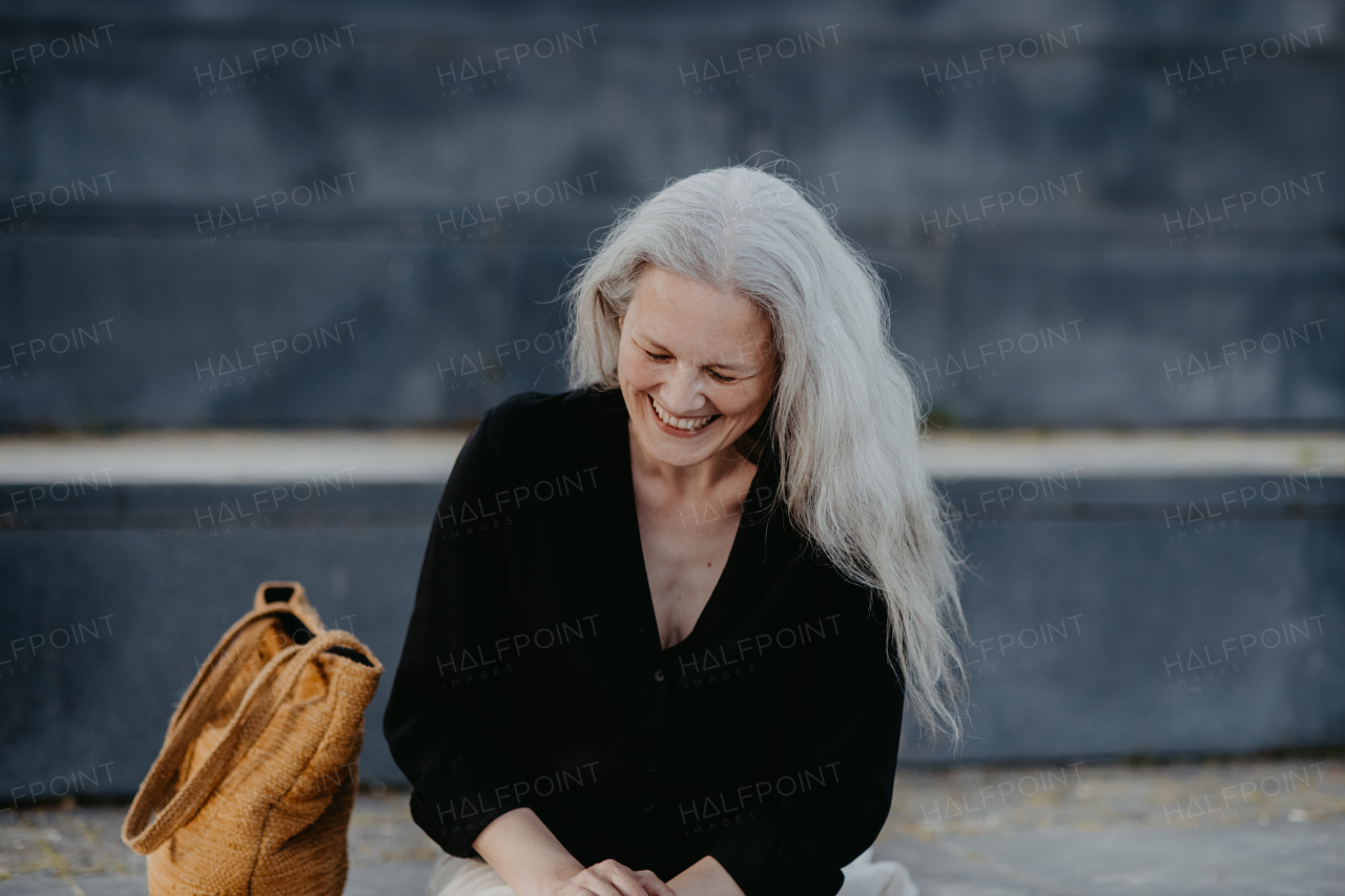 Portrait of a beautiful laughing woman in middle age with long gray hair, standing outdoor in the city.