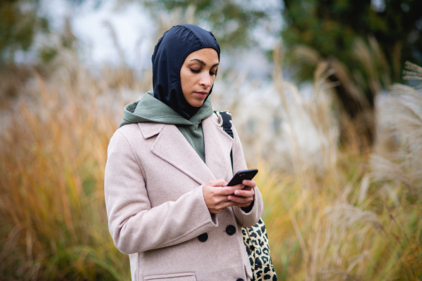 Portrait of young muslim woman in coat, standing outdoor in a city park, scrolling smartphone.