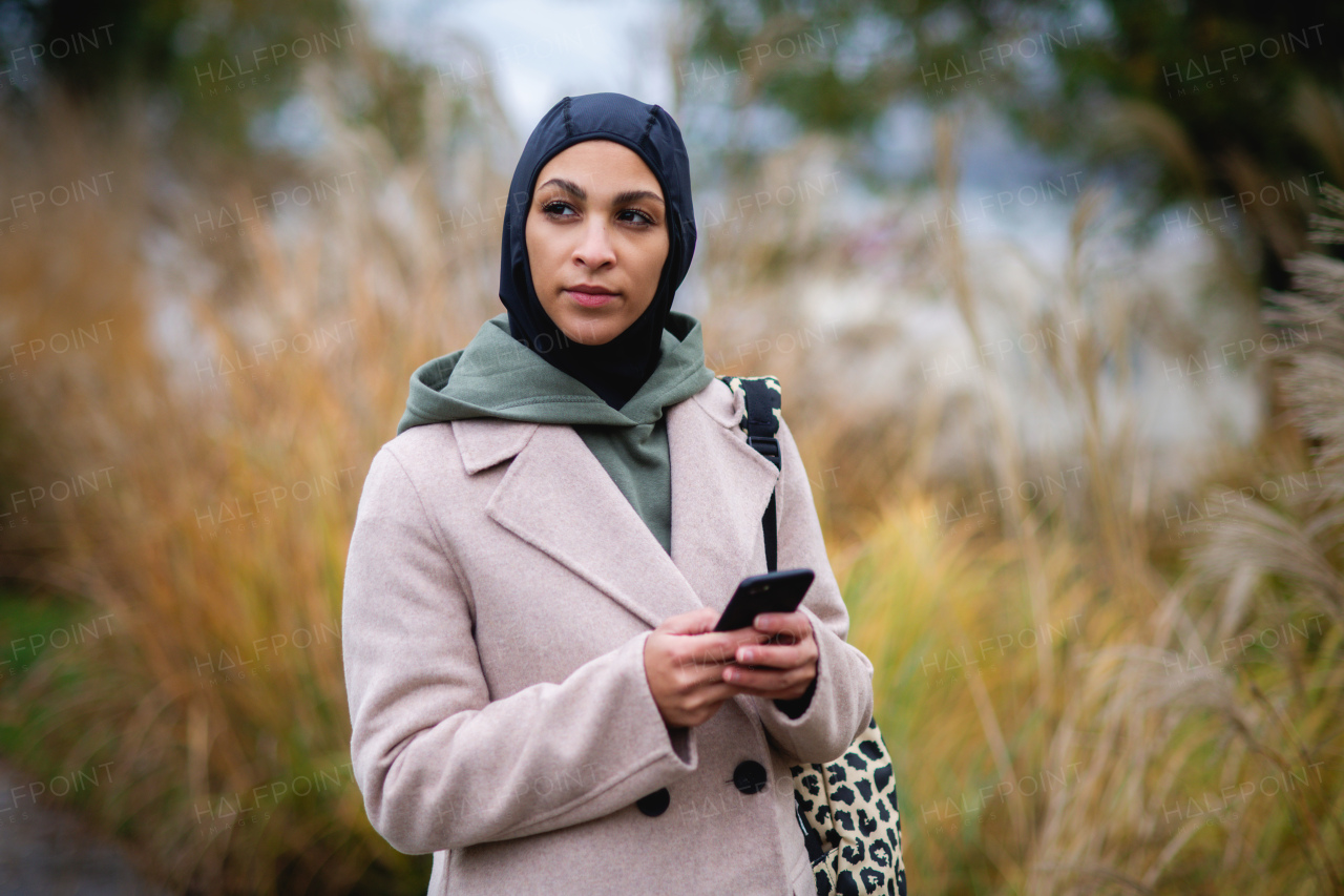 Portrait of young muslim woman in coat, standing outdoor in a city park, scrolling smartphone.