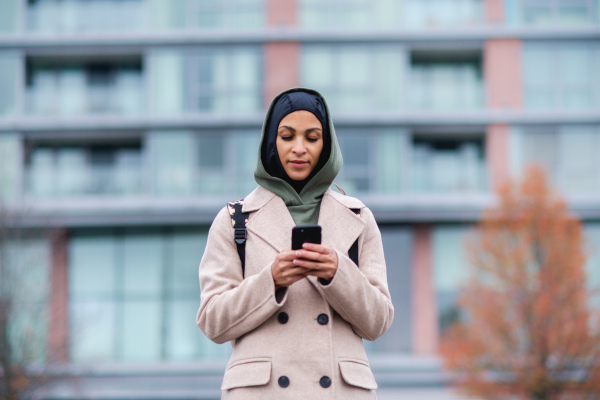 Portrait of young muslim woman in coat, standing outdoor in a city, scrolling smartphone.