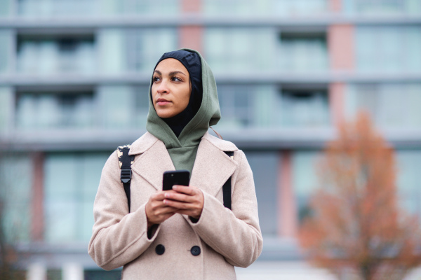 Portrait of young muslim woman in coat, standing outdoor in a city, scrolling smartphone.