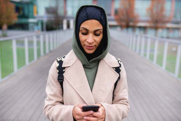 Portrait of young muslim woman in coat, standing outdoor in a city, scrolling smartphone.