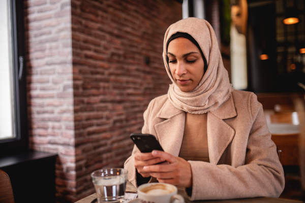 Young muslim woman sitting in cafe and scrolling a smartphone.