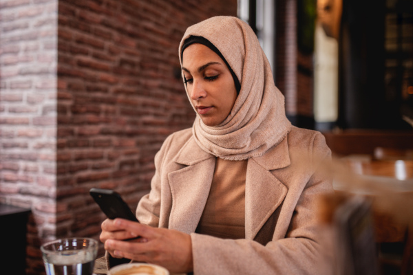 Young muslim woman sitting in cafe and scrolling a smartphone.