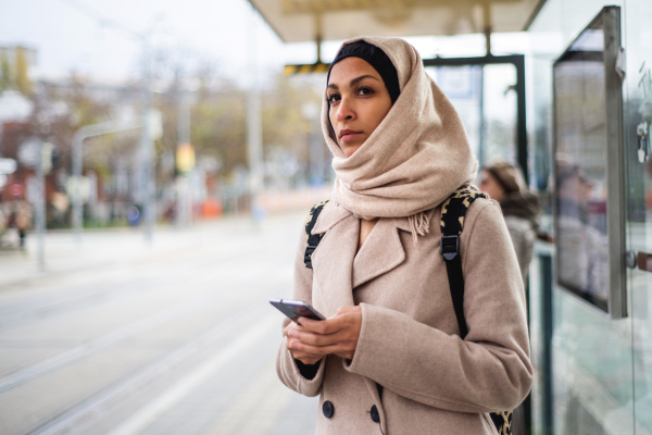 Young muslim woman with a smartphone waiting for bus at city bus stop.