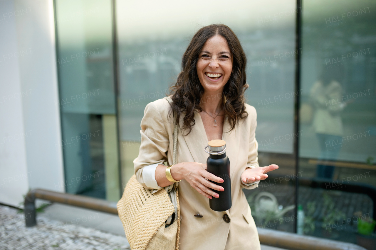 A cheerful woman walking going to work, holding water bottle. Successful happy businesswoman commuting in the morning in city street.