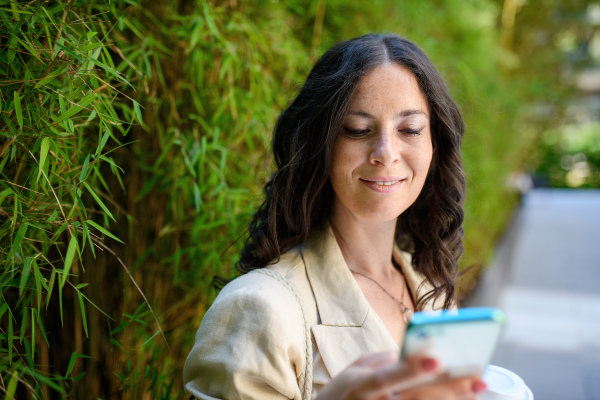 A happy woman traveller, texting on mobile phone and holding cofee cup, with green plant at background.