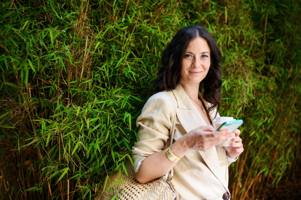 A happy woman traveller, texting on mobile phone and holding cofee cup, with green plant at background.