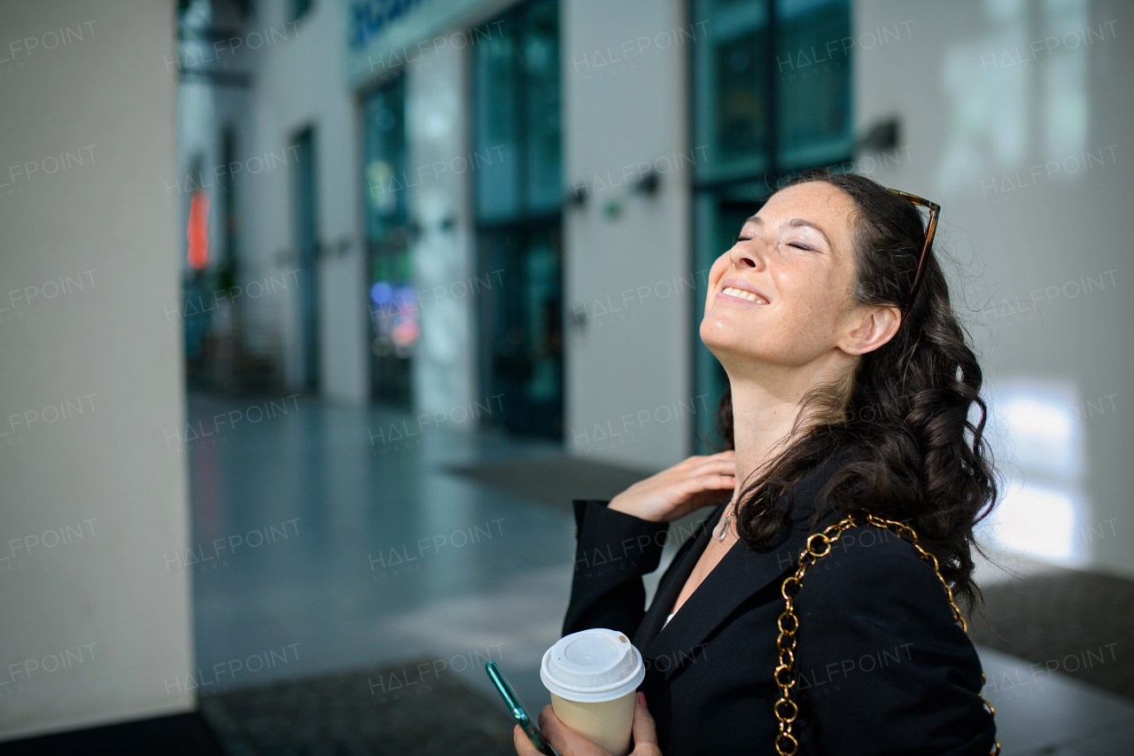 A successful happy businesswoman commuting in the morning in city street, holding cofee cup and sunbathing.