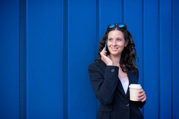 A successful happy businesswoman commuting in the morning in city street, calling on mobile phone and holding cofee cup.