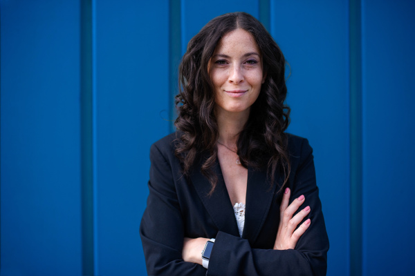 A portriat of happy successful businesswoman commuter with arms crossed standing against blue wall outside in street.