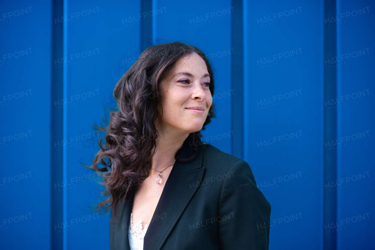 A portriat of happy successful businesswoman commuter with arms crossed standing against blue wall outside in street.