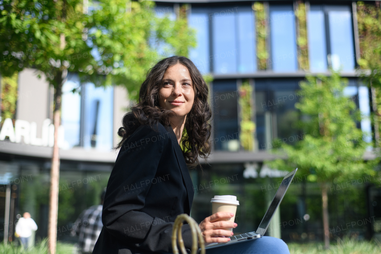 A successful happy businesswomanwith coffe cup sitting and using laptop outside the office buliding.