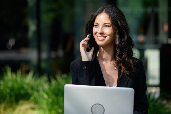 A successful happy businesswoman sitting using laptop and calling on mobile outside the office buliding.