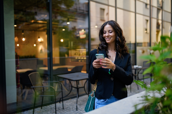 A happy woman texting on mobile phone and waiting outside the cafe in city.