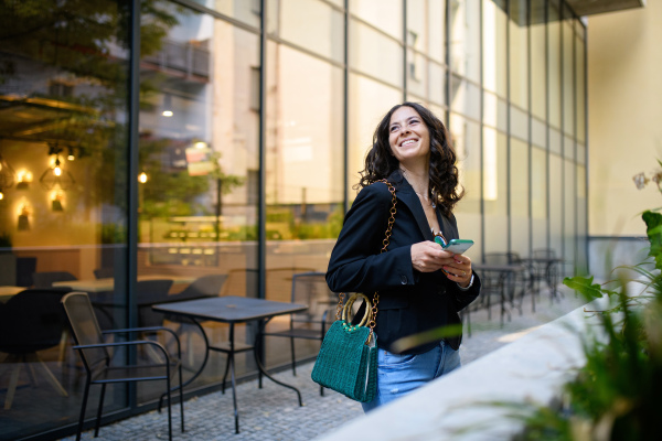 A happy woman texting on mobile phone and waiting outside the cafe in city.