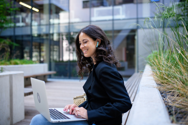 A successful happy businesswoman sitting and using laptop outside the office buliding.