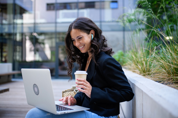 A successful happy businesswoman sitting and using laptop outside the office buliding.