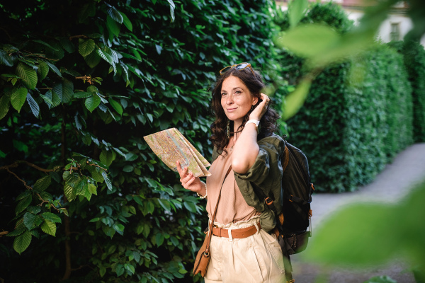 A young beautiful female traveler with map standing in park and looking in map on sunny day in city.