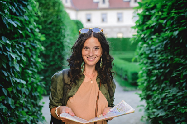 A young beautiful female traveler with map standing in park and looking in map on sunny day in city.