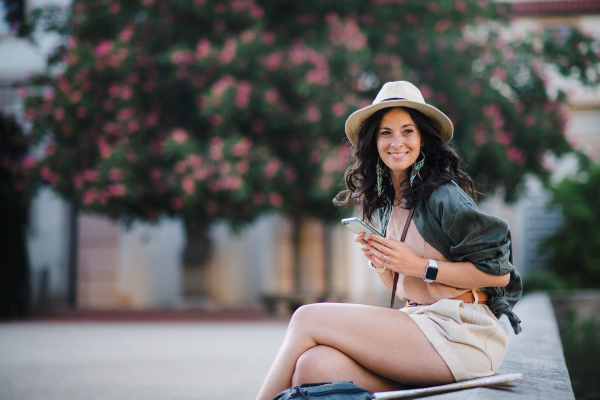 A young beautiful female traveler with cellphone sitting in park near pond on sunny day in city.