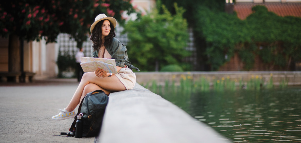 A young beautiful female traveler with map sitting in city park near pond on sunny day in city.