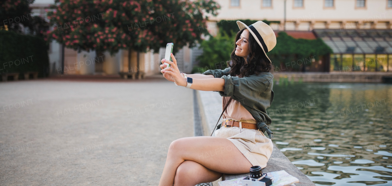 A young beautiful female traveler sitting on bridge and taking selfie, on sunny day in city.