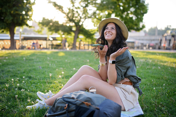 Young beautiful female sitting in a park and taking selfie on sunny day in city.