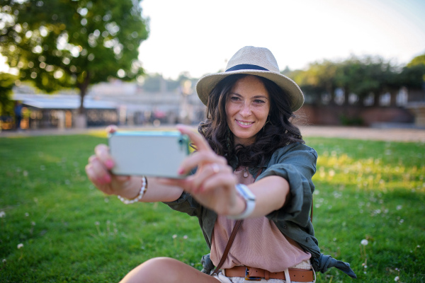 Young beautiful female sitting in a park and taking selfie on sunny day in city.