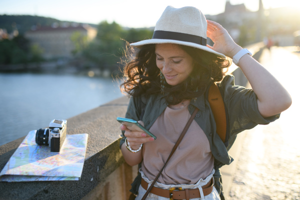 A young beautiful female traveler with map standing on bridge and using cellphone on sunny day in city.