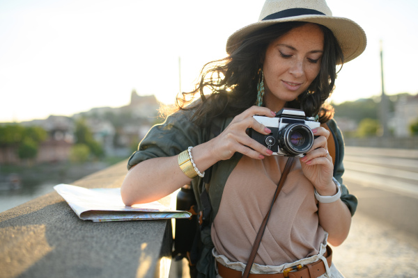 A young beautiful female traveler with map standing on bridge and using camera on sunny day in city.