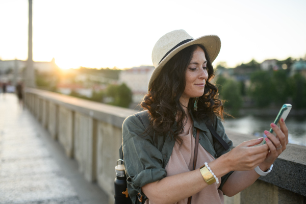 A young beautiful female traveler with backpack walking through bridge and taking selfie, on sunny day in city.