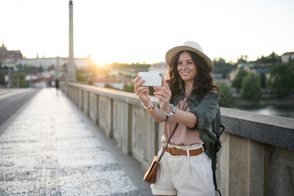 A young beautiful female traveler with backpack walking through bridge and taking selfie, on sunny day in city.