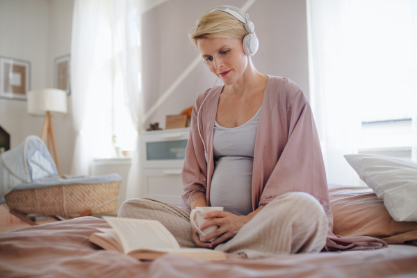 Pregnant woman sitting in a bed, listening music and reading book, enjoying time for herself.