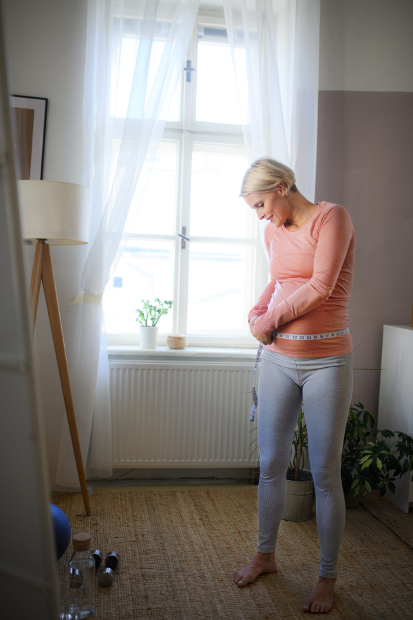 Happy woman measuring her pregnant belly in her apartment.