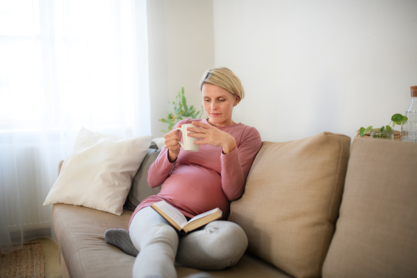 Pregnant woman sitting on sofa, reading book and enjoying a cup of tea.