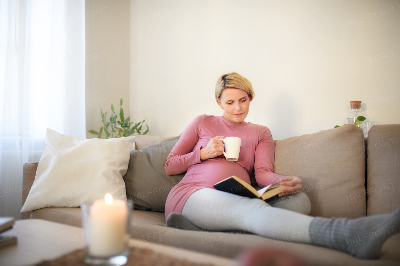 Pregnant woman sitting on sofa, reading book and enjoying a cup of tea.